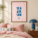 Sea Air Sun – Blauwe Letters op Roze Strepen in een lichte slaapkamer met crème wand, roze linnen beddengoed, blauwe lamp en walnootkleurige lijst