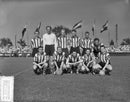 Het elftal van PSV op het veld bij de wedstrijd tegen Elinkwijk op 23 september 1956, met vlaggen en publiek op de achtergrond.