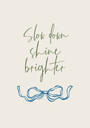 Groene handgeschreven Slow down shine brighter letters boven een blauwe lintstrik op crème.
